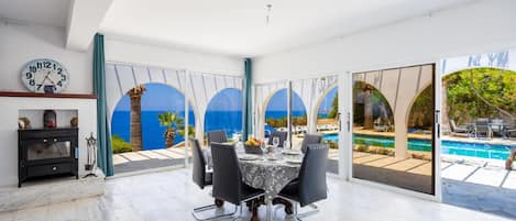 Une salle à manger moderne avec une table dressée pour quatre personnes, de grandes fenêtres donnant sur une piscine et des palmiers, et une horloge au mur.