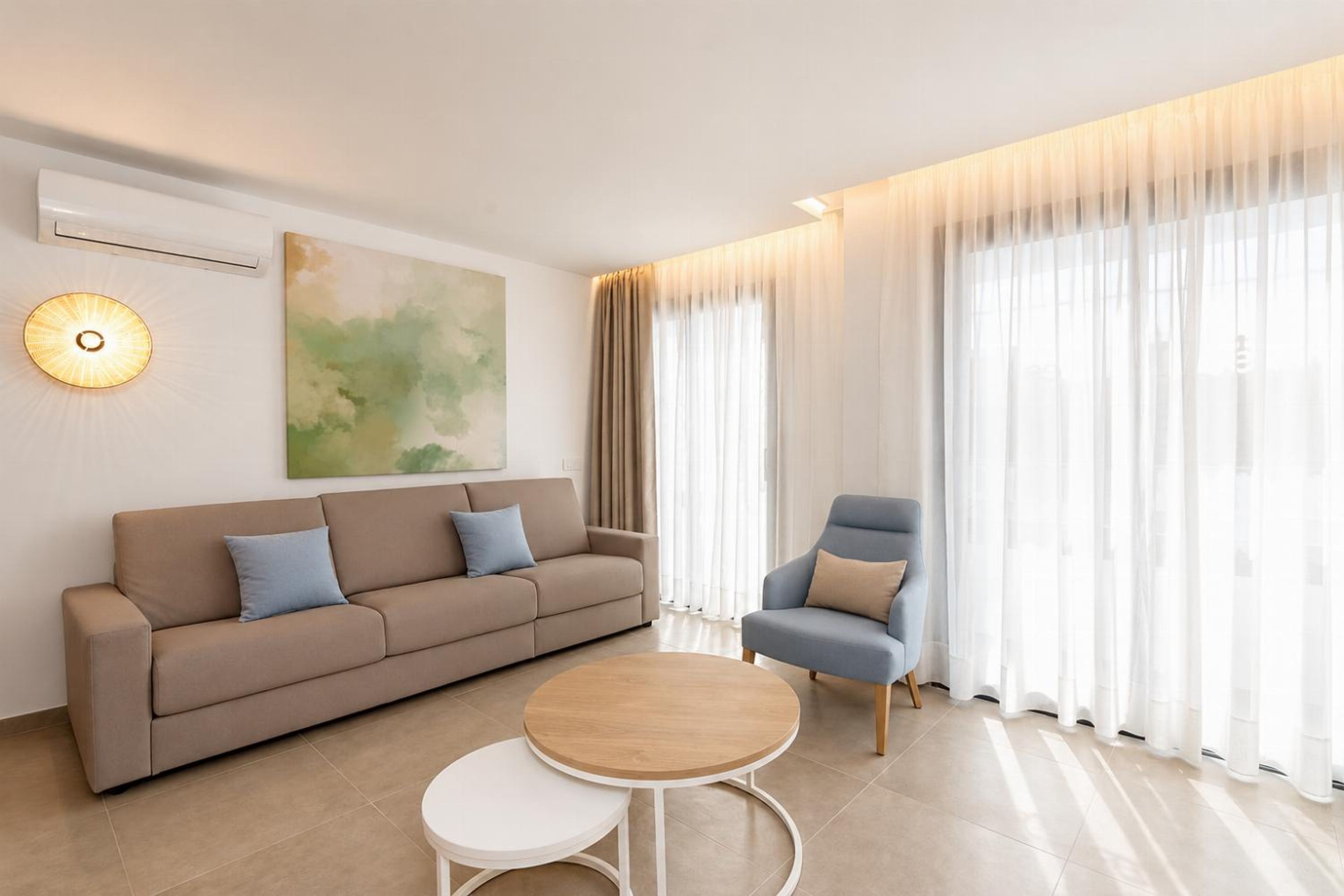 Una sala de estar moderna con un sofá, un sillón, una mesa de centro redonda y un ventanal con cortinas traslúcidas.