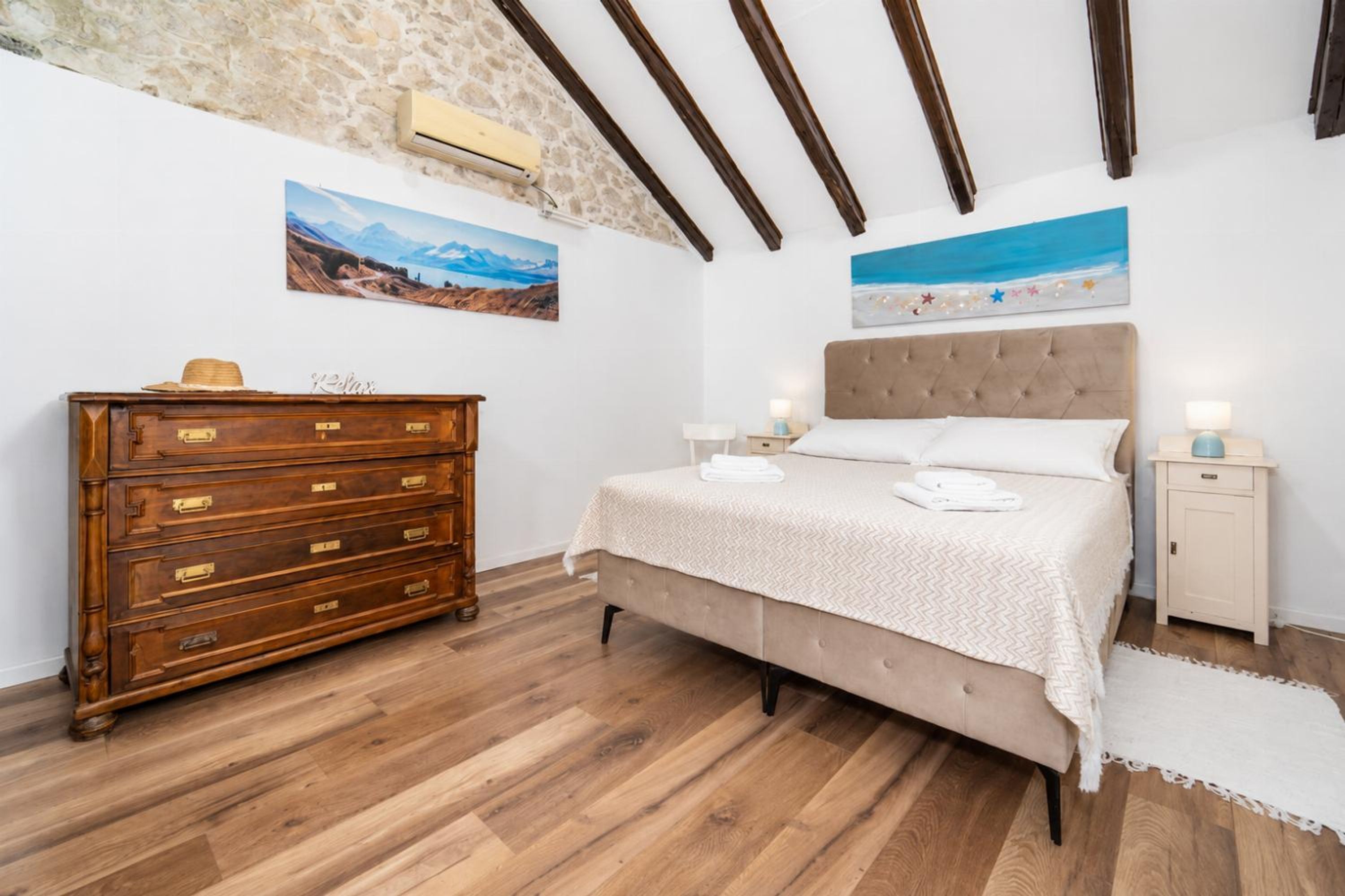 A bedroom with a wooden dresser, a bed with a beige headboard, and two paintings on the walls.