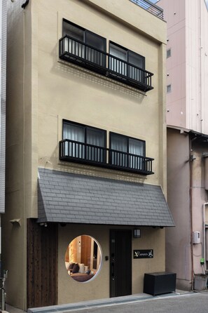 Ein Gebäude mit einer beigefarbenen Fassade, zwei Balkonen mit schwarzen Geländern und einem Rundfenster. Ein Schild trägt die Aufschrift 'Yamazaki'.