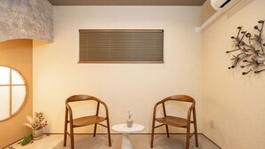 Ein Zimmer mit zwei Holzsesseln um einen kleinen weißen Tisch, ein Fenster mit Jalousien, einer Wand-Klimaanlage und einem dekorativen Metallkunstwerk.