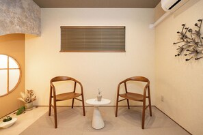 Ein Zimmer mit zwei Holzsesseln um einen kleinen weißen Tisch, ein Fenster mit Jalousien, einer Wand-Klimaanlage und einem dekorativen Metallkunstwerk.