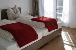 Ein Bett mit einer karierten Decke und roten Tagesdecken, ein Nachttisch mit Lampe sowie der Blick auf einen Balkon durch ein Fenster.