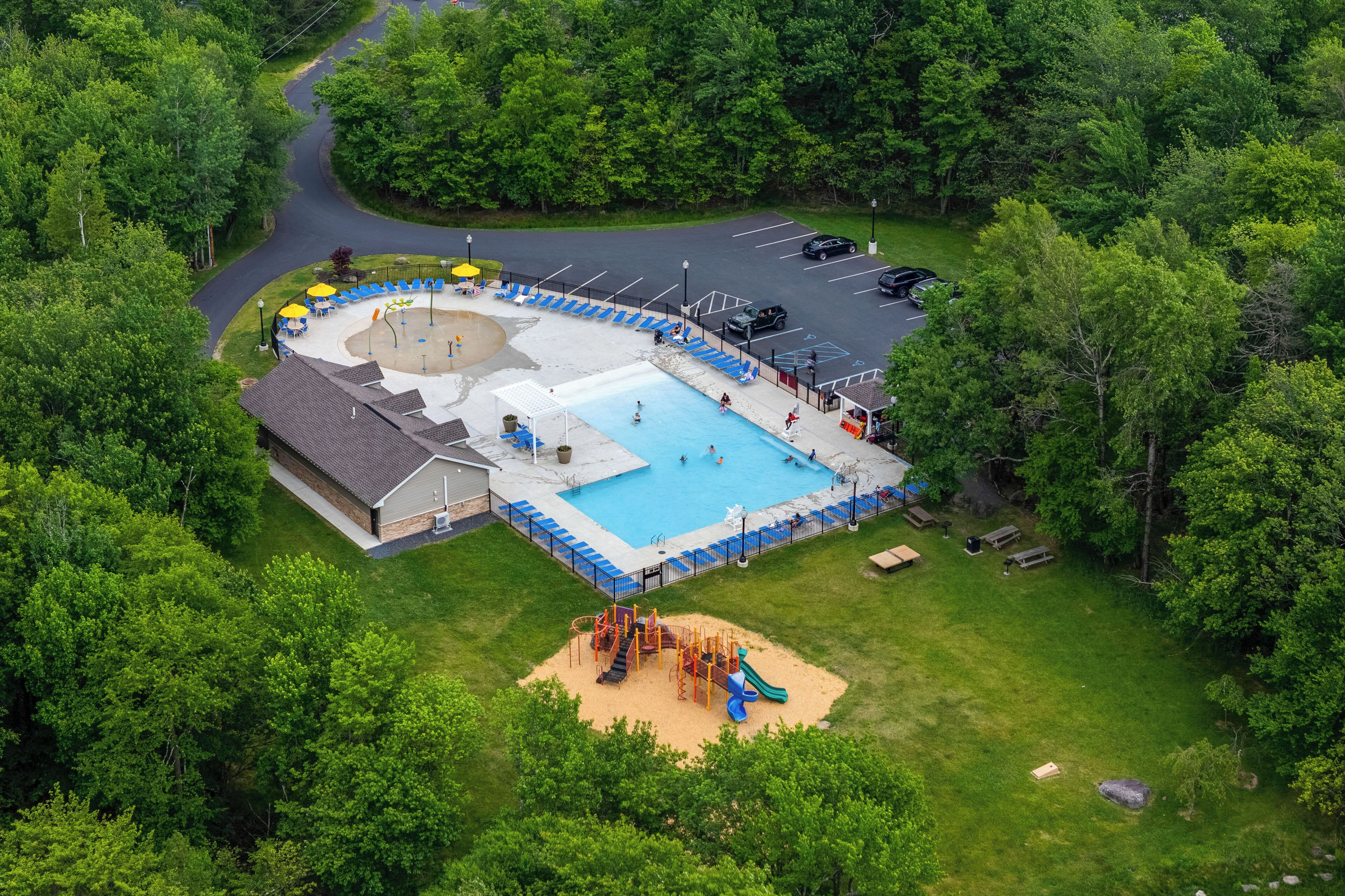 Vue aérienne d’un espace piscine comprenant une aire de jeux, un espace de repos couvert et un parking.