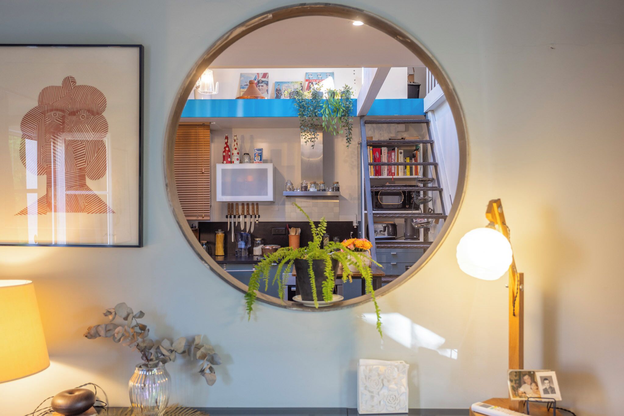 A round mirror reflecting a kitchen area with a bookshelf, a plant, and a framed picture on the wall.