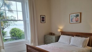 Ein Schlafzimmer mit einem hölzernen Bett, einem Nachttisch, einer Lampe, einem Fenster mit Vorhängen und einem gerahmten Bild an der Wand.