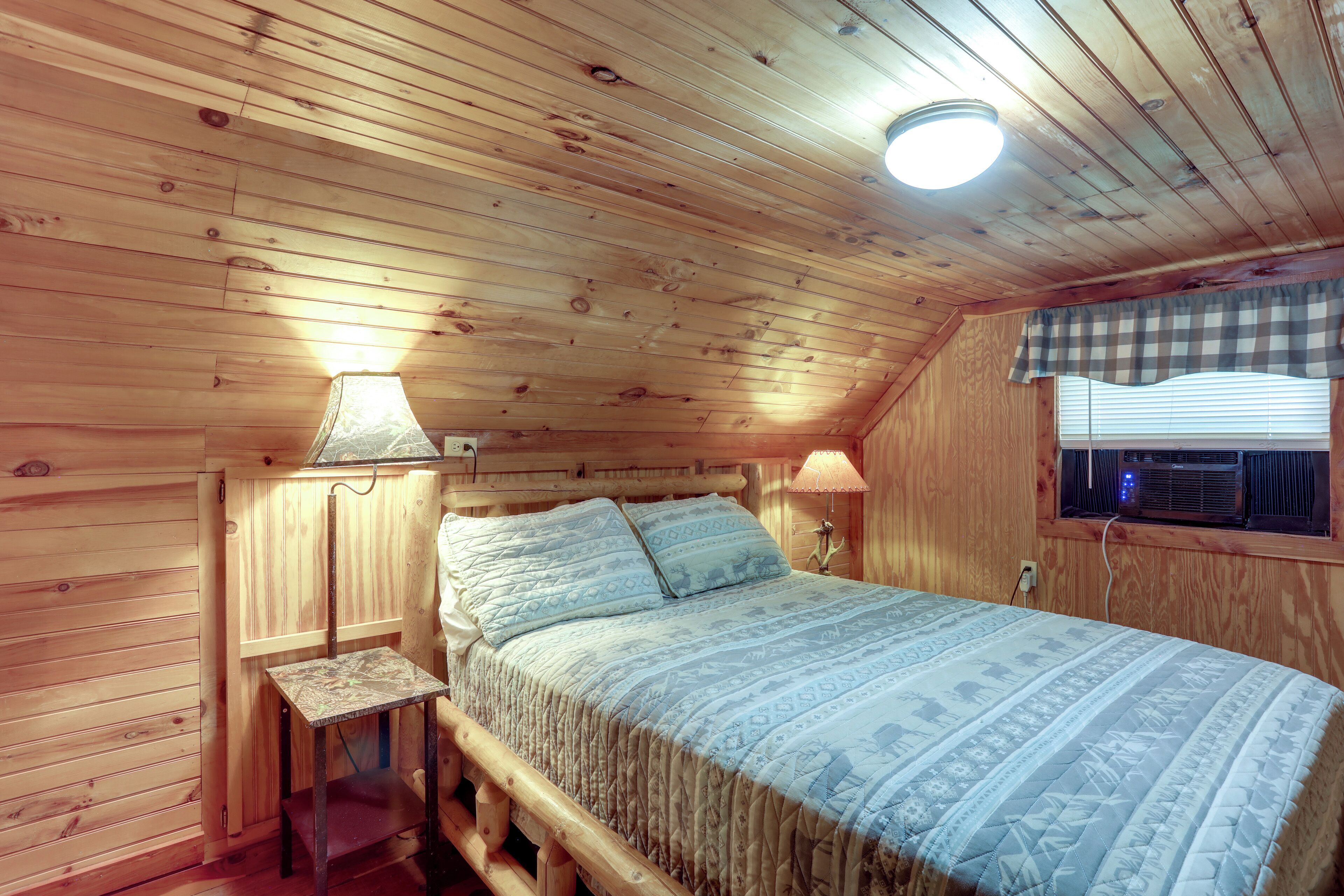 Un dormitorio de cabaña de madera con cama, dos lámparas, una ventana con persianas y una lámpara de techo.
