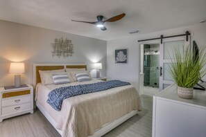 Ein Schlafzimmer mit Bett, Nachttischen, Deckenventilator, einer Topfpflanze und einer Schiebetür zum Badezimmer.