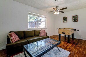 A living room with a glass coffee table, a sofa with striped pillows, a wooden foosball table, a ceiling fan, and a window with blinds. - Suburban Sanctuary: Magnolia Home w/ Backyard! (Magnolia)