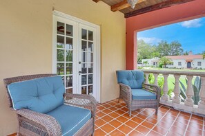 Eine überdachte Terrasse mit zwei Korbstühlen, einer weißen Tür und Blick auf ein Haus im Hintergrund.