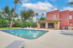 Ein Swimmingpool mit gefliestem Rand, eine Liegefläche mit Lounge-Stühlen und ein Gebäude mit roter Fassade und Ziegeldach.