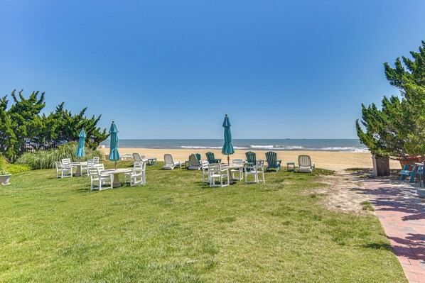 Eine begrünte Fläche mit Sitzgelegenheiten und Sonnenschirmen, ein Weg zum Strand und Blick auf das Meer.