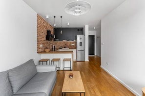 Ein modernes Wohnzimmer mit einem grauen Sofa, einem Holztisch und einer Küchenzeile mit Backsteinwand, Barhocker und einem Kühlschrank.