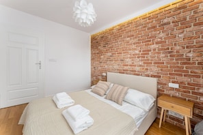 A bedroom with a brick wall, a bed with white linens, a wooden nightstand, and a white door. - Riverside Residence • 1BR • Elevator • Old Town (Kraków)