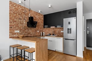 A modern kitchen with a brick wall, white cabinets, a stainless steel refrigerator, and bar stools. - Riverside Residence • 1BR • Elevator • Old Town (Kraków)
