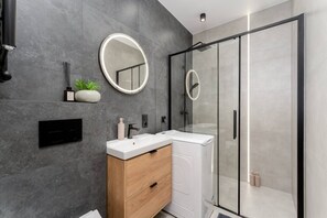 A modern bathroom with a glass shower enclosure, a round mirror, a wooden vanity, and a built-in washing machine. - A4 • Couples Retreat | Old Town 5 min (Kraków)