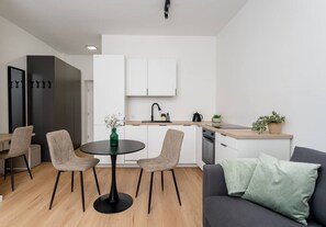 Eine moderne Küche mit weißen Schränken, ein Essbereich mit einem runden Tisch und Stühlen sowie ein graues Sofa mit einem grünen Kissen.
