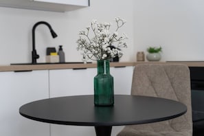 Ein runder Tisch mit einem grünen Blumenvasen voller weißer Blüten, eine Küchenzeile mit einem schwarzen Wasserhahn und ein beiger Stuhl.