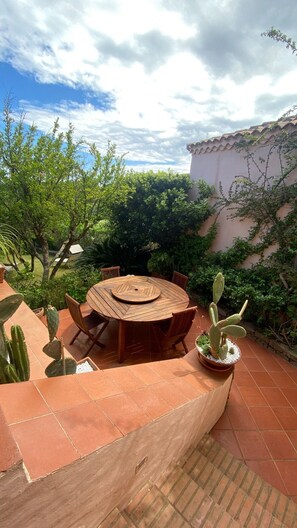 Une table ronde en bois, entourée de chaises, de cactus et de plantes, est placée sur une terrasse carrelée, avec un bâtiment visible en arrière-plan.