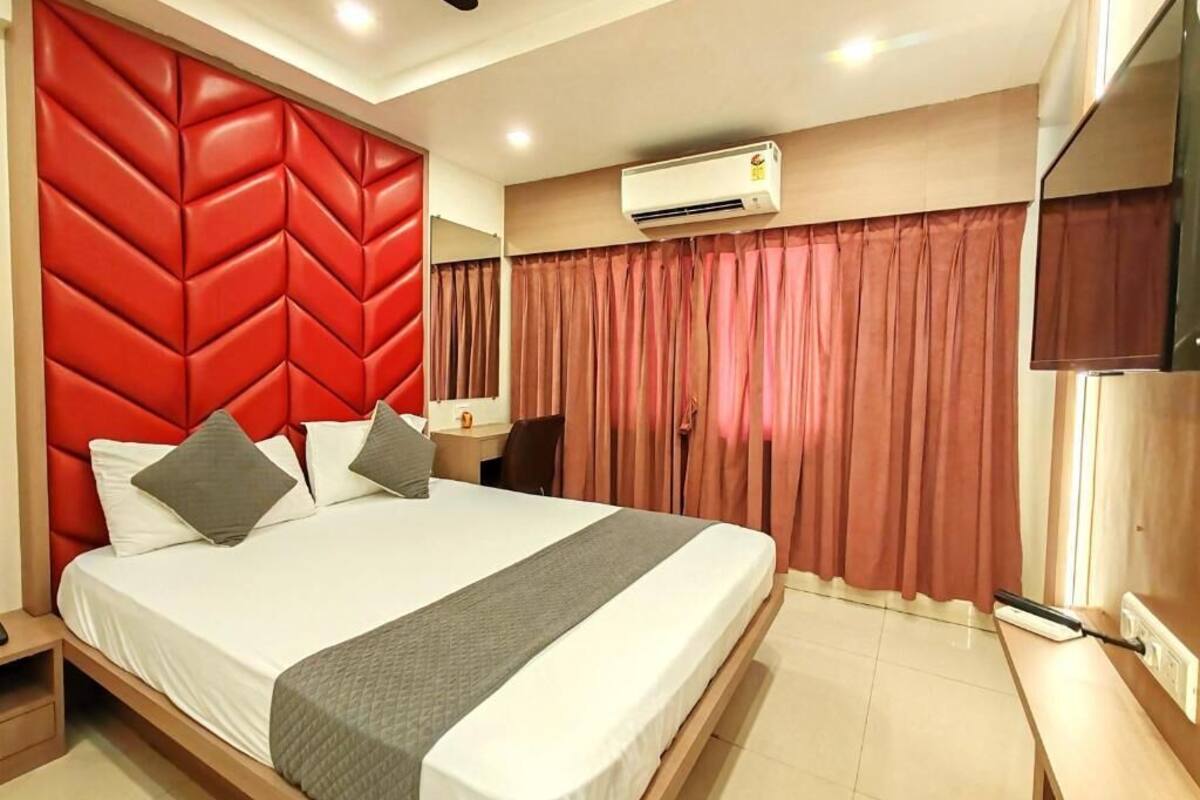 A hotel room with a large bed, a red headboard, an air conditioning unit, a desk, and a mirror.
