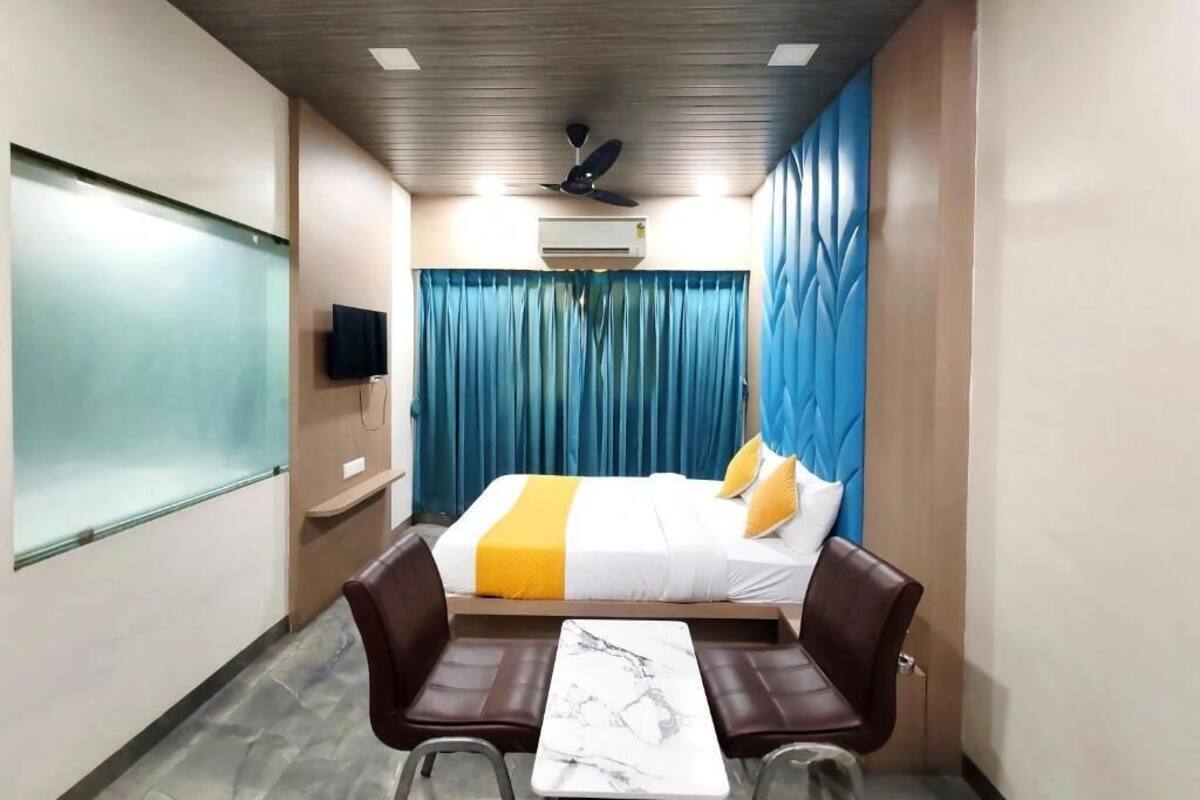 A hotel room with a bed, a desk, two chairs, a ceiling fan, an air conditioning unit, and a large window with curtains.