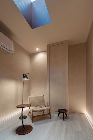 Um quarto com clarabóia no teto, luminária de chão, cadeira de vime, um pequeno banquinho e um ar condicionado fixado na parede.