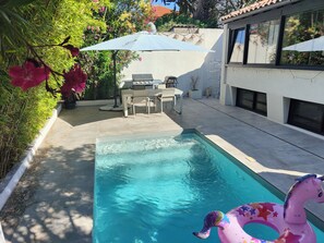 A swimming pool with a pink inflatable unicorn float, a patio area with a table and chairs, and a covered seating area with a mural. - Modern House in Marseille's 9th District, Mazargues, 10 minutes from the sea and 20 minutes from Cassis. (Marseille)