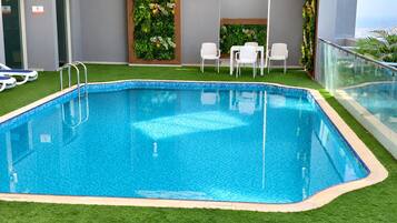 Une piscine rectangulaire intérieure à l’eau d’un bleu cristallin, entourée de gazon artificiel, de chaises longues blanches et d’une rambarde en verre.