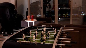 Une table de baby-foot dans une pièce dotée d’une grande fenêtre, une petite table avec une boisson et une chaise.