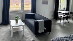 Ein modernes Wohnzimmer mit einer Couch, einem kleinen Tisch mit Pflanze, einer Essecke mit Tisch und Stühlen sowie großen Fenstern mit Vorhängen.