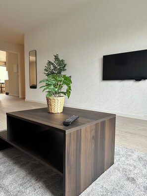A modern living room with a dark wood coffee table, a potted plant, a remote control, a flat-screen TV, and a woven basket.