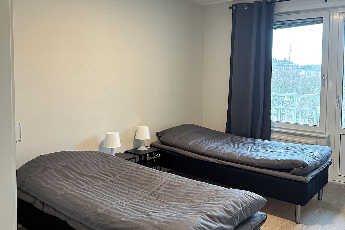 A bedroom with two beds, a window with curtains, a bedside table with lamps, and a view of a cityscape.