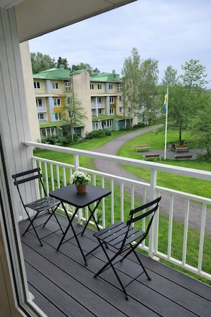 A balcony with a table and chairs, overlooking a green lawn and a building with multiple windows.