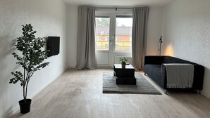 Ein Wohnzimmer mit einer Couch, einem Couchtisch mit Pflanze, einem Fernseher an der Wand und Blick durch ein Fenster.