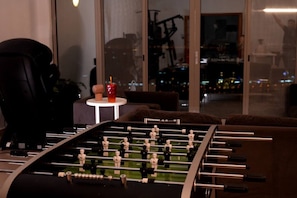 A foosball table in a room with a large window, a small table with a drink, and a chair.