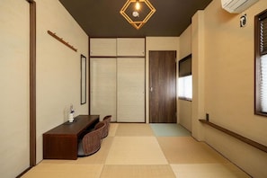 Une pièce de style japonais traditionnel, avec un sol recouvert de tatamis, une basse table en bois entourée de chaises, une porte coulissante et un luminaire au plafond.