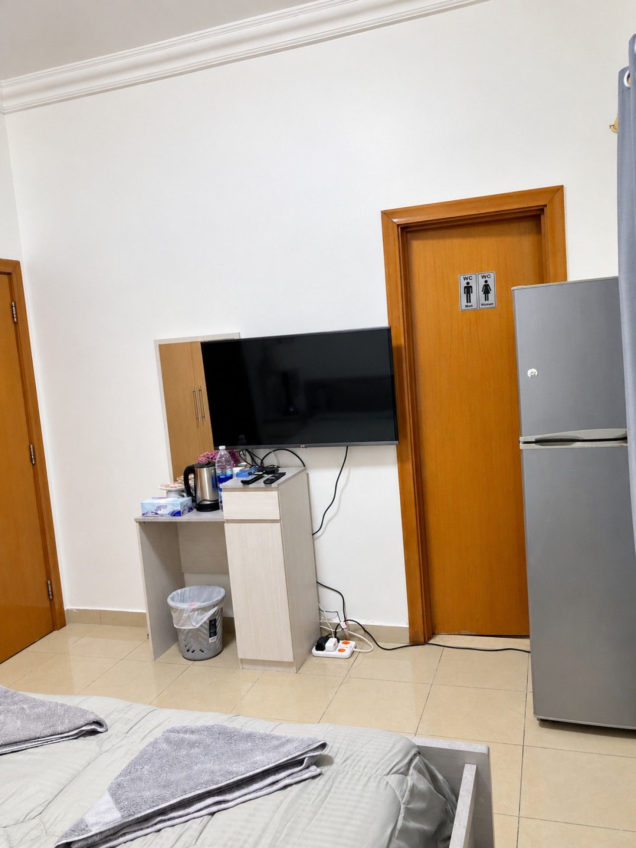 Quarto de hotel compacto com uma cama, TV fixada na parede, pequeno espaço de cozinha com geladeira e uma lixeira.