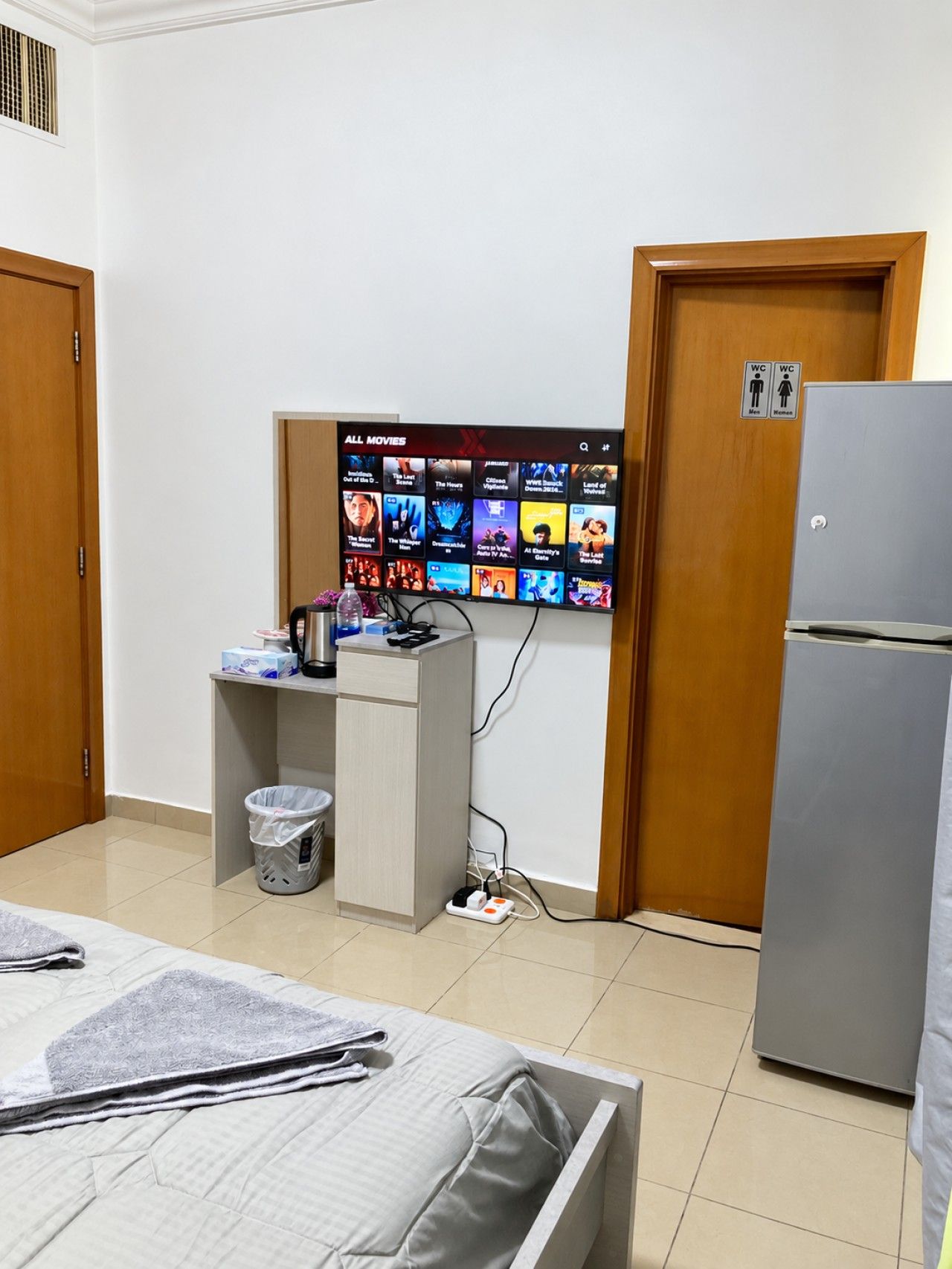 Ein Hotelzimmer mit einem Bett, einem Schreibtisch mit Computer, einem an der Wand befestigten Fernseher der Filmoptionen zeigt, einem Mülleimer und einem Kühlschrank.