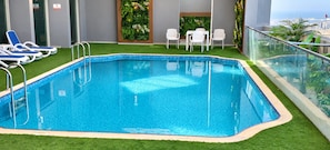 A rectangular indoor pool with clear blue water, surrounded by artificial grass, white lounge chairs, and a glass railing.
