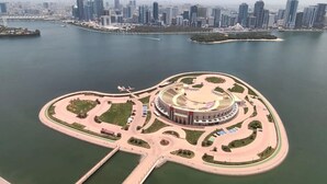 An aerial view of a waterfront property with a circular central structure, a marina, and a city skyline in the background.