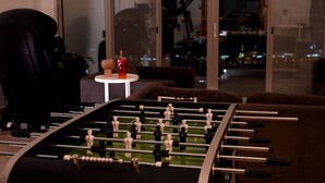 A foosball table in a room with a large window, a small table with a red cup, and a chair.
