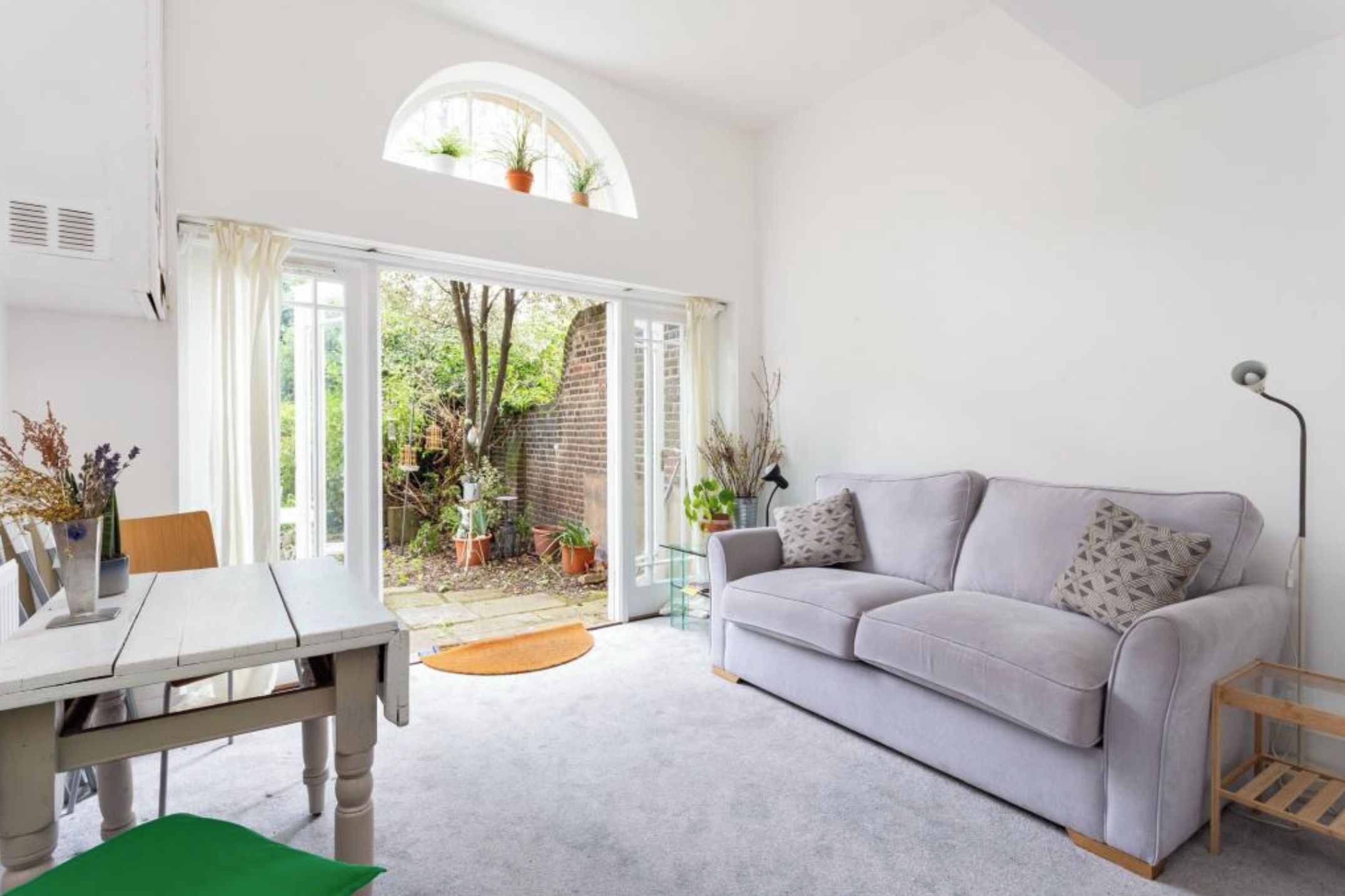 A bright living room with a white sofa, a dining table, and a view of a garden through a glass door.