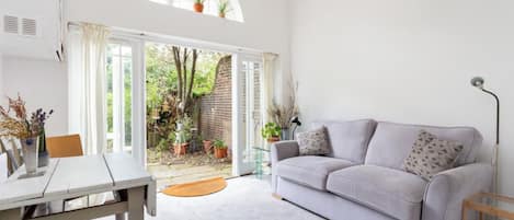 A bright living room with a white sofa, a dining table, and a view of a garden through a glass door.