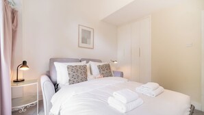 Ein ordentlich bezogenes Bett mit weißen Bettwäsche und Handtüchern, ein Nachttisch mit Lampe sowie ein Kleiderschrank.