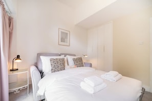 A neatly made bed with white linens and towels, a bedside table with a lamp, and a wardrobe. - Bright 1BDR Flat in the heart of Little Portugal (Greater London)