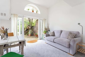 A living room with a sofa, a table, and a window overlooking a garden. - Charming 1BDR Flat in Stockwell (Greater London)