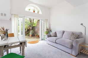 A bright living room with a white sofa, a dining table, and a view of a garden through a glass door. - Bright 1BDR Flat in the heart of Little Portugal (Greater London)