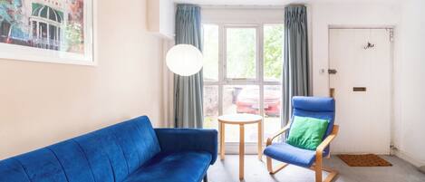 Une pièce avec un canapé bleu, une chaise en bois avec un coussin vert, une petite table ronde et une porte blanche.
