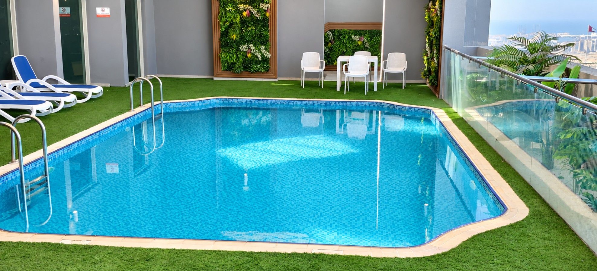 A rectangular indoor pool with clear blue water, surrounded by artificial grass, white lounge chairs, and a glass railing.