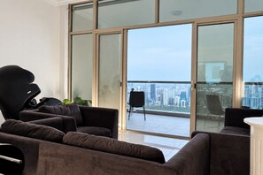 Een moderne woonkamer met een massagestoel, een bank en een stadsgezicht door grote glazen deuren.